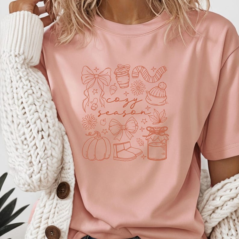 May include: A light pink t-shirt featuring a collection of fall-themed illustrations in a burnt orange color. The design includes bows, a coffee cup, a scarf, a beanie, a pumpkin, and the words "cozy season".