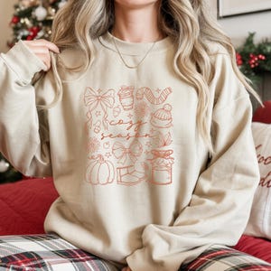 May include: Beige sweatshirt with a red graphic that says "cozy season" and features drawings of bows, a coffee cup, a scarf, a hat, flowers, a pumpkin, and a jar. The sweatshirt is a neutral color.