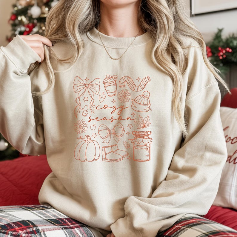 May include: Beige sweatshirt with a red graphic that says "cozy season" and features drawings of bows, a coffee cup, a scarf, a hat, flowers, a pumpkin, and a jar. The sweatshirt is a neutral color.
