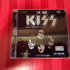 May include: A clear plastic CD jewel case featuring a black cover with the band name KISS in large white letters. The artwork includes four band members in makeup, the text THE OMNI, OCTOBER 1, 1996, ATLANTA, GA, ALIVE WORLDWIDE, and 2025 NSU RECORDS.