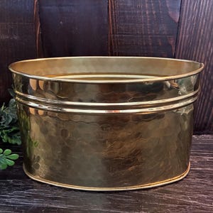 Puede incluir: Una maceta ovalada de latón con un acabado pulido y martillado. El recipiente presenta un borde redondeado y liso, junto con una banda decorativa horizontal que rodea la parte superior del cuerpo, mostrando una superficie metálica dorada.