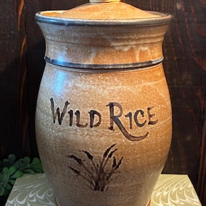 May include: A tan ceramic canister with a lid, featuring the text "WILD RICE" in dark brown lettering above a painted illustration of wild rice stalks. The vessel has a speckled texture, a flared rim, and a dark brown band around the top edge.