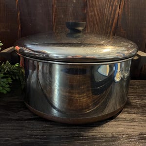 May include: A stainless steel cooking pot with a polished reflective finish, featuring a matching lid with a central black knob and two black side handles. The base of the pot has a distinct copper-coloured band around the bottom edge.