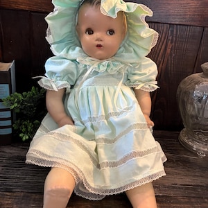 May include: A vintage doll with painted facial features, wearing a light mint green dress with puffed sleeves and a matching bonnet. The dress and bonnet are decorated with white lace trim and ribbons. The doll has a smooth, light-toned complexion.