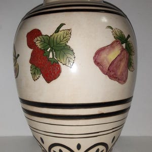 May include: Cream-colored ceramic vase with black stripes and fruit illustrations. The vase features red strawberries, purple pears, and green leaves. The base has a decorative black and blue floral design.