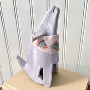 May include: A ceramic dolphin sculpture in a light purple colour. It features a pink band with colourful triangle designs. The sculpture is placed on a beige surface against a white background.
