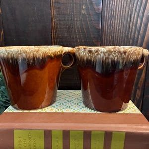 Pode incluir: Duas canecas de cerâmica com acabamento brilhante, apresentando uma base castanho-escura que transita para um padrão de gotejamento em tom creme claro na borda. Cada caneca possui uma alça resistente e uma abertura larga e alargada.