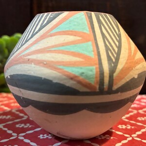 May include: A small, round, matte-finish ceramic vase featuring a geometric pattern in shades of teal, terracotta, and charcoal gray. The upper portion displays angular lines and leaf-like shapes, while the lower section has a scalloped border.