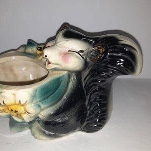 May include: A vintage ceramic skunk planter with a whimsical design. The skunk has black and white fur, pink cheeks, and gold accents. The planter is shaped like a flower and has a teal and yellow color scheme. A unique decorative piece.