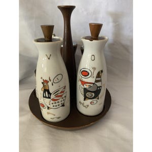 May include: A vintage condiment set featuring two white ceramic bottles with wooden stoppers and whimsical bird illustrations. The set includes a tall, dark brown wooden handle and sits on a round, dark brown wooden tray.