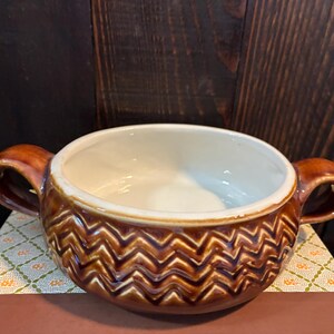 May include: A round ceramic soup bowl featuring two side handles and a glossy brown exterior with a raised, textured chevron pattern. The interior of the bowl is a smooth, solid off-white colour, providing a contrast to the dark brown outer surface.