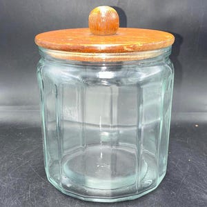 May include: A clear glass storage jar with a hexagonal body and a flat, circular wooden lid. The lid features a small, rounded wooden knob handle centered on top. The glass container has vertical panels and a wide opening for storage purposes.