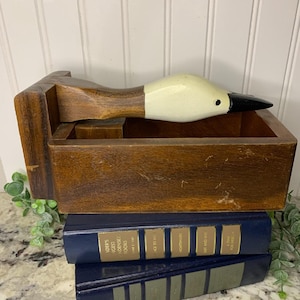Op de afbeelding: Een houten eendvormige doos met een witte en zwarte eendenkop, rustend op twee marineblauwe boeken met gouden letters. De doos is warmbruin van kleur en heeft een rechthoekige vorm.