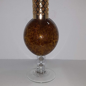 May include: A decorative glass fragrance lamp with a gold-colored metal top and a round, speckled amber-colored glass body. The lamp sits on a clear glass stem and base, creating an elegant and stylish design.