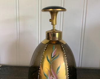 Puede incluir: Un frasco de perfume antiguo con una tapa de metal dorado y una tapa negra. La botella de vidrio tiene un tono marrón oscuro con un panel dorado con un diseño floral en rosa y verde. Puntos blancos bordean el panel dorado.