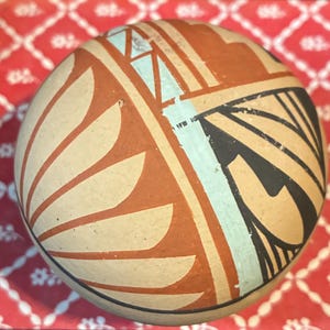 May include: A spherical ceramic vessel featuring a matte tan base color decorated with hand-painted geometric patterns in terracotta orange, black, and pale blue. The design includes repeating leaf-like shapes and linear segments.