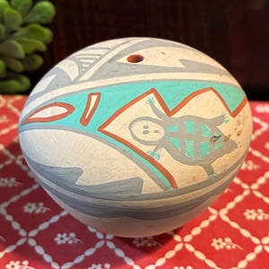 May include: A spherical ceramic vessel with a matte off-white surface featuring hand-painted geometric patterns in turquoise, grey, and terracotta. A stylized turtle figure is depicted on the side, and a small circular opening is at the top.