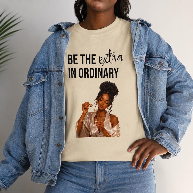 May include: A beige t-shirt featuring the black text BE THE extra IN ORDINARY above a graphic of a person with dark curly hair in a high bun wearing a white satin robe and holding a small glass dropper bottle, layered under a blue denim jacket.