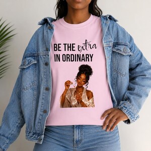 May include: A light pink t-shirt featuring the black text BE THE extra IN ORDINARY above a graphic of a person with dark hair in an updo wearing a white robe and holding a small glass dropper bottle, layered under a blue denim jacket.