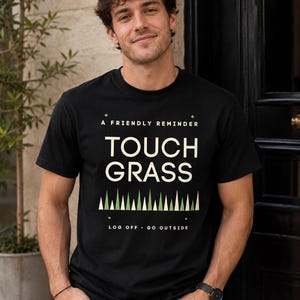 Op de afbeelding: Een zwart t-shirt met korte mouwen met de tekst A FRIENDLY REMINDER TOUCH GRASS in cr&egrave;me, een rij groene en cr&egrave;mekleurige driehoeken eronder, en de tekst LOG OFF GO OUTSIDE onderaan in een strak, gecentreerd grafisch ontwerp.