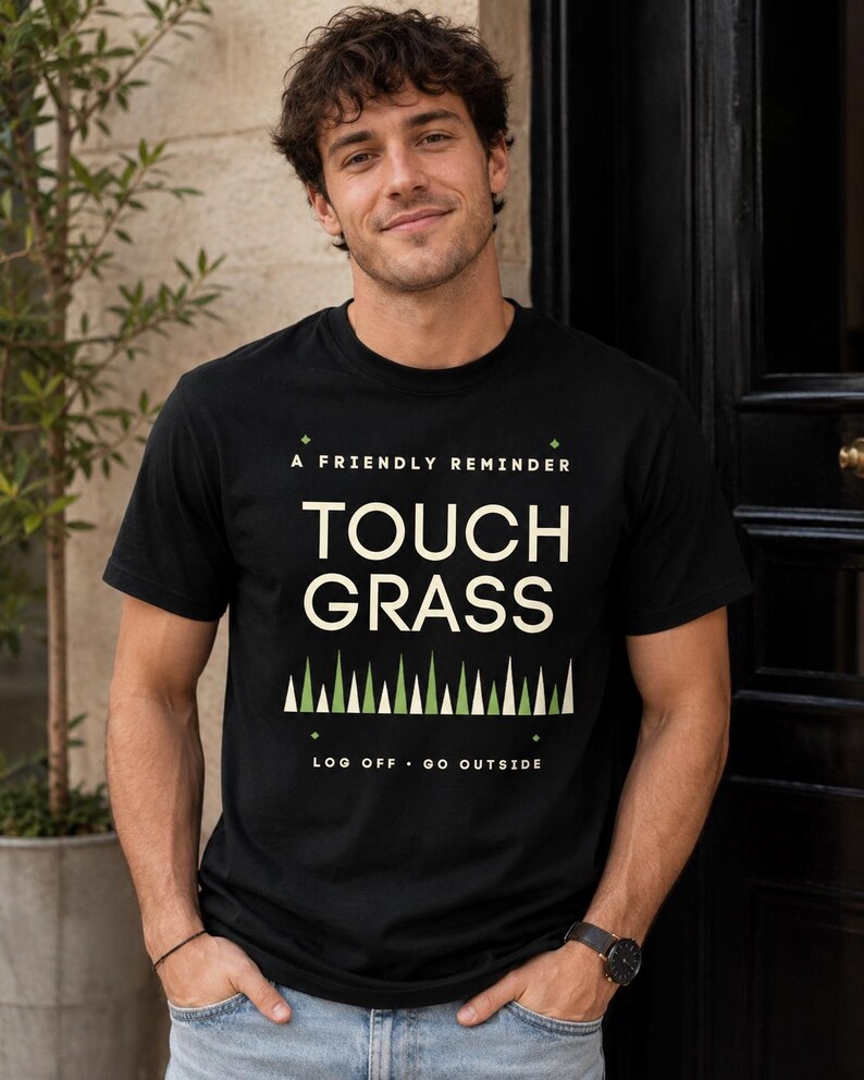 Op de afbeelding: Een zwart t-shirt met korte mouwen met de tekst A FRIENDLY REMINDER TOUCH GRASS in cr&egrave;me, een rij groene en cr&egrave;mekleurige driehoeken eronder, en de tekst LOG OFF GO OUTSIDE onderaan in een strak, gecentreerd grafisch ontwerp.