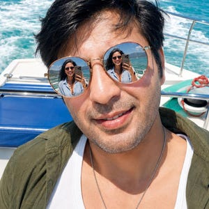Puede incluir: Gafas de sol redondas con montura de metal fino en tono dorado y lentes reflectantes. Las lentes muestran el reflejo claro de una persona sosteniendo un tel&eacute;fono inteligente. Las gafas cuentan con almohadillas ajustables y patillas finas.
