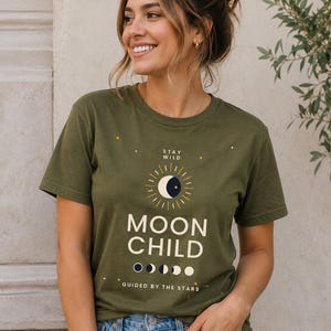 Op de afbeelding: Een olijfgroen t-shirt met korte mouwen en een grafische print met de tekst "STAY WILD", "MOON CHILD" en "GUIDED BY THE STARS". Het ontwerp bevat een zon- en maanicoon, een reeks maanfasen en kleine witte en gele steraccenten.