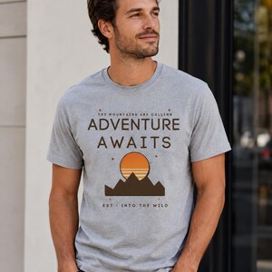 Op de afbeelding: Een grijs gem&ecirc;leerd t-shirt met korte mouwen en een grafische print met de tekst "THE MOUNTAINS ARE CALLING ADVENTURE AWAITS" en "EST. INTO THE WILD" boven en onder een gestileerd bruin gebergte en een ronde oranje zon.