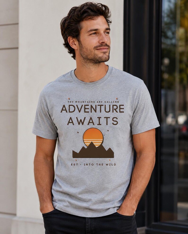 Op de afbeelding: Een grijs gem&ecirc;leerd t-shirt met korte mouwen en een grafische print met de tekst "THE MOUNTAINS ARE CALLING ADVENTURE AWAITS" en "EST. INTO THE WILD" boven en onder een gestileerd bruin gebergte en een ronde oranje zon.