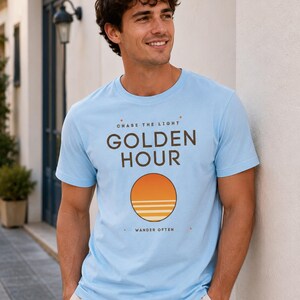 Op de afbeelding: Een lichtblauw t-shirt met korte mouwen en de tekst CHASE THE LIGHT, GOLDEN HOUR en WANDER OFTEN in donkerbruine letters. Onder de tekst staat een cirkelvormige afbeelding van een oranje zon die ondergaat boven horizontale witte lijnen.