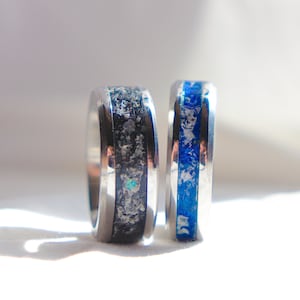 May include: Two metal rings standing upright. The left ring features a wide black inlay with silver-toned flecks and a small iridescent accent. The right ring features a vibrant blue inlay with silver-toned flecks throughout the band.