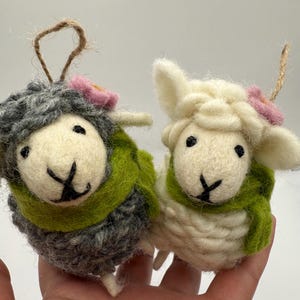 May include: Two handmade sheep ornaments. One is gray, the other is white, both with black eyes and a stitched black mouth. Each sheep wears a green scarf and a pink flower. They have a jute string for hanging.