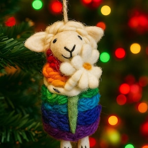 May include: A handcrafted sheep ornament with a rainbow-colored body and a white daisy. The sheep has a cream-colored head and is suspended by a natural twine string. The background is a blurred Christmas tree with colorful lights.