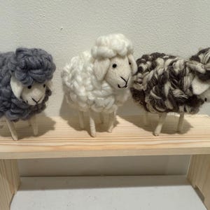 Puede incluir: Tres figuras de ovejas de lana en patrones grises, blancos y marrones y blancos, de pie sobre una pequeña estantería de madera. Cada oveja tiene un cuerpo de lana esponjosa, cara blanca y finas patas de madera. Las ovejas son hechas a mano y decorativas.