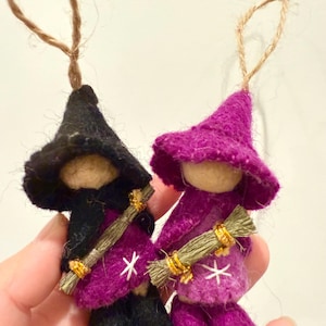 May include: Two handmade witch ornaments. One witch is wearing a black hat and outfit, the other a purple hat and outfit. Both have a broom and a white star on their outfit. Each ornament has a jute string for hanging.