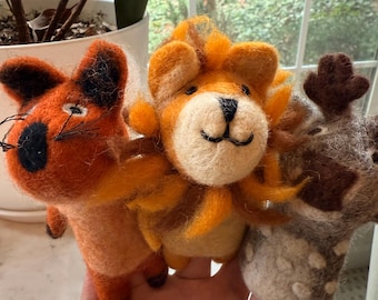 May include: Three handmade felt animal figurines: an orange fox, a lion with a yellow mane, and a giraffe with brown spots. The fox has black whiskers and a black eye. The lion has a smiling face. The giraffe has brown antlers. These are likely children's toys.