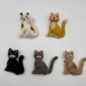 Könnte beinhalten: Fünf handgefertigte Katzenfiguren aus Filz in verschiedenen Farben. Die Katzen sind weiß mit orangefarbenen Flecken, gelb mit weißen Akzenten, schwarz, grau und beige mit braunen Streifen. Jede Katze hat einen einzigartigen Ausdruck und eine einzigartige Pose.