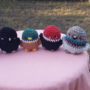 May include: Five handmade crochet bird figurines in various colors, including yellow, black, teal, red, and gray. The birds are arranged on a pink surface, with a grassy background.