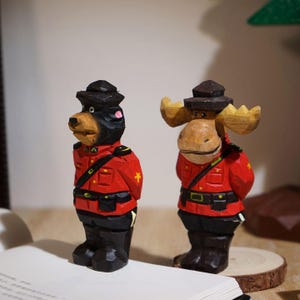 Könnte beinhalten: Zwei handgeschnitzte Holzfiguren: ein Bär und ein Elch, beide in roten Jacken, schwarzen Hosen und Hüten gekleidet, die an Mounties erinnern. Der Bär hat einen schwarzen Kopf und der Elch hat große Hörner. Die Figuren stehen aufrecht.