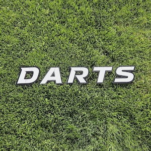 Könnte beinhalten: Weißes und schwarzes "DARTS"-Schild mit schwarzer Umrandung, das auf einer grünen Grasfläche liegt. Die fetten Buchstaben sind ideal für ein Spielzimmer oder eine Sportdekoration.