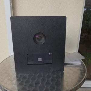 Op de afbeelding: Een zwart, vierkant apparaat met een lens en een klein rechthoekig paneel met het nummer "50". Het apparaat staat op een ronde, zilveren tafel.