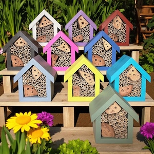 May include: A collection of colorful wooden bee houses in various shades, including blue, pink, and yellow. Each house features a small, triangular roof and is filled with bamboo tubes and wooden inserts, designed to attract bees. The houses are arranged on a wooden surface.