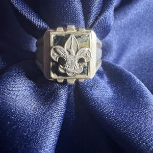 May include: A silver-toned ring with a rectangular face featuring a fleur-de-lis emblem. The emblem is set against a dark background. The ring is displayed on a blue fabric.