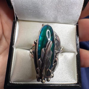 May include: A silver ring featuring an elongated, iridescent abalone shell inlay with a deep green and blue color palette. The shell is framed by detailed, sculpted silver leaves that wrap around the sides of the central stone setting.