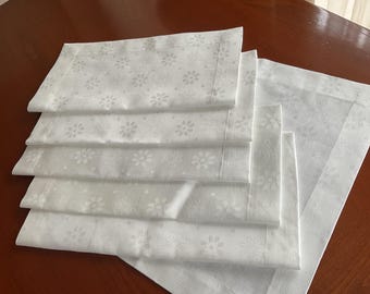 May include: A stack of white cloth napkins with a floral pattern and a decorative border. The napkins are neatly folded and arranged on a wooden surface, suggesting a dining or table setting theme.