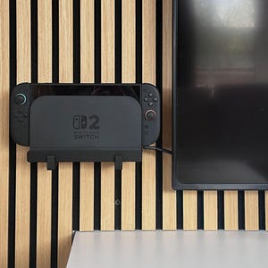 Könnte beinhalten: Eine schwarze Nintendo Switch Konsole, die mit einer schwarzen Kunststoffhalterung an der Wand befestigt ist. Die Konsole verfügt über zwei abnehmbare Controller mit einem blauen Ring am linken Joystick und einem roten Ring am rechten Joystick.