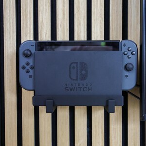 Könnte beinhalten: Eine dunkelgraue Nintendo Switch-Konsole in einer Dockingstation mit grauen Joy-Con-Controllern. Die Dockingstation ist an einer Holzwand mit vertikalen schwarz-beigen Streifen befestigt. Das Nintendo Switch-Logo ist sichtbar.