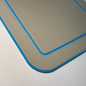 May include: A rectangular, light gray mat with a blue border. The mat has a textured surface and a blue design in the upper corner. The edges are rounded, and the mat is set against a white background.