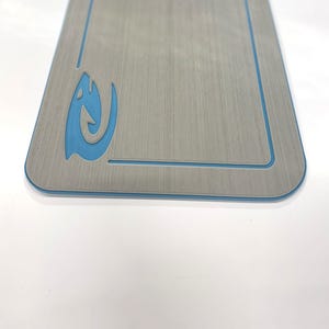 Puede incluir: Una tabla rectangular de madera gris con un borde azul y un dise&ntilde;o de pez estilizado azul en la esquina superior izquierda. La tabla tiene una textura de vetas de madera y esquinas redondeadas.