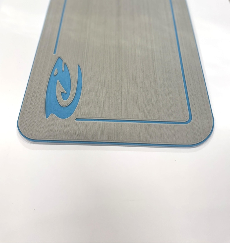 Puede incluir: Una tabla rectangular de madera gris con un borde azul y un dise&ntilde;o de pez estilizado azul en la esquina superior izquierda. La tabla tiene una textura de vetas de madera y esquinas redondeadas.